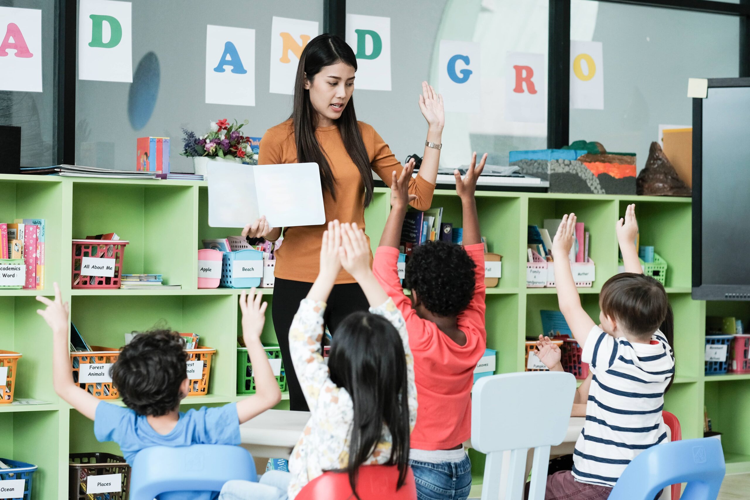 Teacher standing in front of young students reading a book. The teacher and the students have their hands raised. The classroom is colorful.