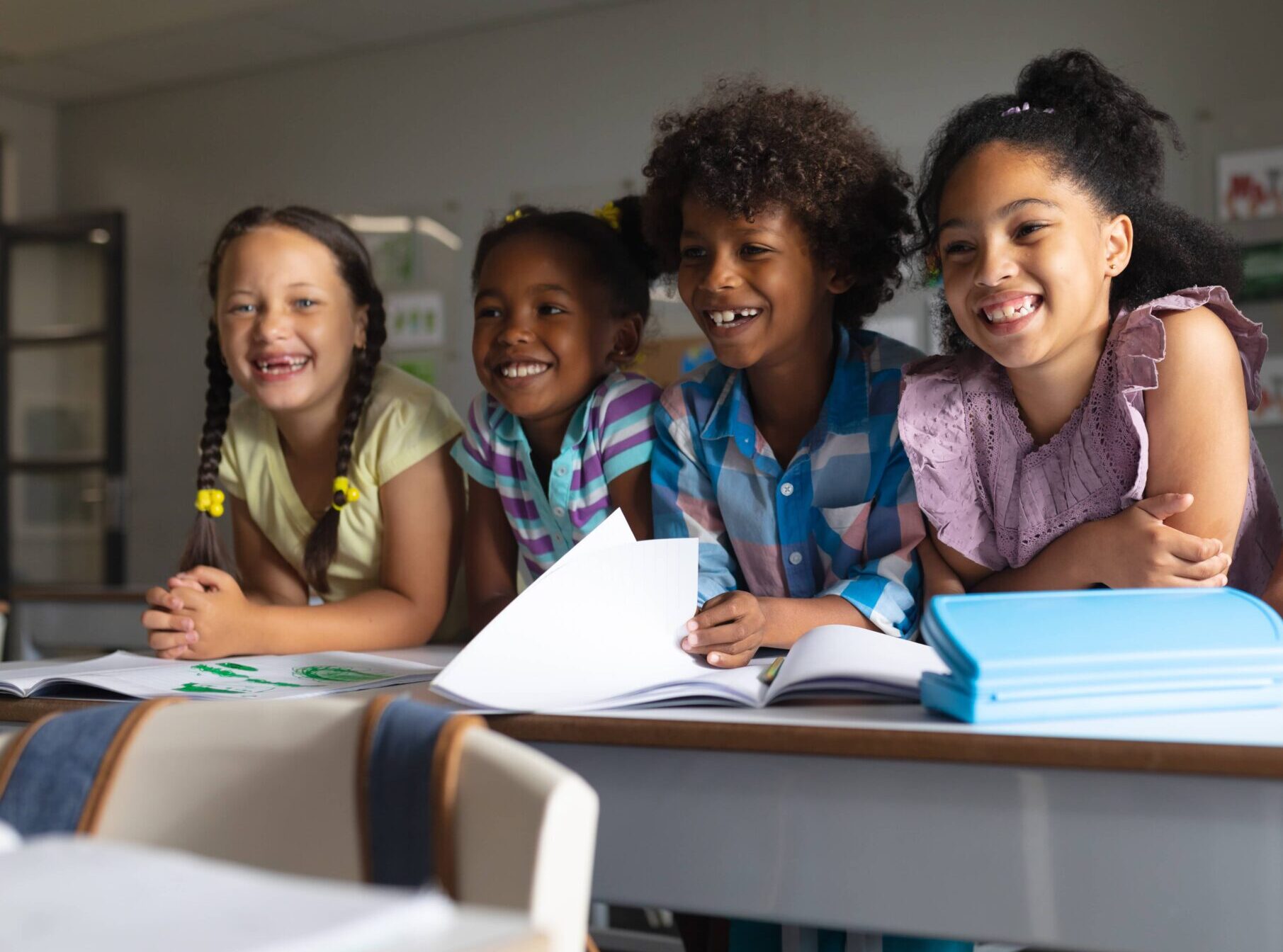 Four students leaning on desks with papers. they are laughing and smiling.